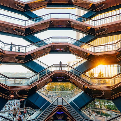 Multi-level stair structure with people walking and standing, framed by a cityscape and sunset reflection on adjacent buildings.