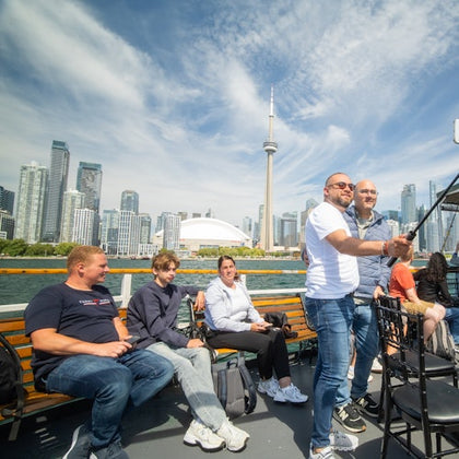 Group of people sitting and standing on a boat with Toronto's skyline, including the CN Tower, visible in the background.