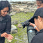 Three people on a rocky, mossy shore observe something small in one person's hand. One holds a camera.