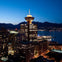 A cityscape at dusk featuring a lit tower, buildings, and a waterfront with mountains in the background.