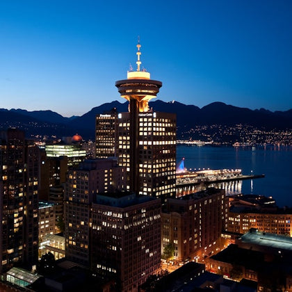 A cityscape at dusk featuring a lit tower, buildings, and a waterfront with mountains in the background.