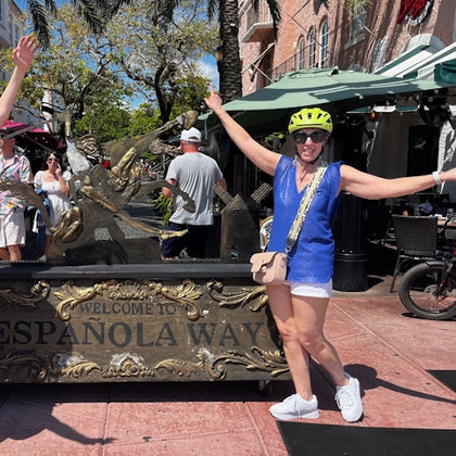Person in a yellow helmet and blue top posing by a "Welcome to Espanola Way" sign, with people and a cafe in the background.