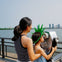 A woman and a child wearing a green crown look through a coin-operated viewer near a waterfront with city buildings in the background.