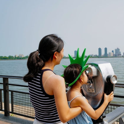 A woman and a child wearing a green crown look through a coin-operated viewer near a waterfront with city buildings in the background.