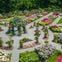 A formal garden with pathways, flowerbeds, and a gazebo covered with flowering vines, surrounded by lush greenery.