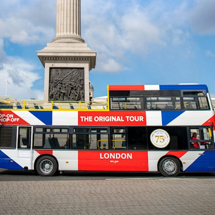 A double-decker bus with a Union Jack design and "The Original Tour" branding, parked near a column monument on a sunny day.