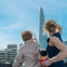 A woman and a child pointing toward the Shard skyscraper in London, with the cityscape and a clear blue sky in the background.