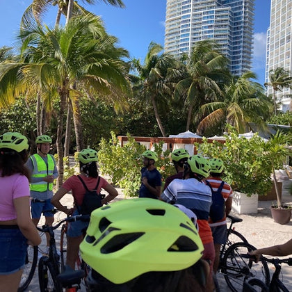 Group of people wearing yellow helmets with bicycles, standing near palm trees and tall buildings, under a clear blue sky.