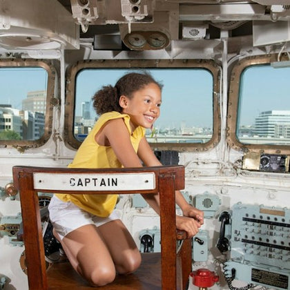 A child sitting in a ship's captain's chair inside a control room with windows showing a cityscape in the background.