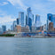 City skyline with modern skyscrapers and waterfront buildings, viewed from across a body of water under a blue sky.