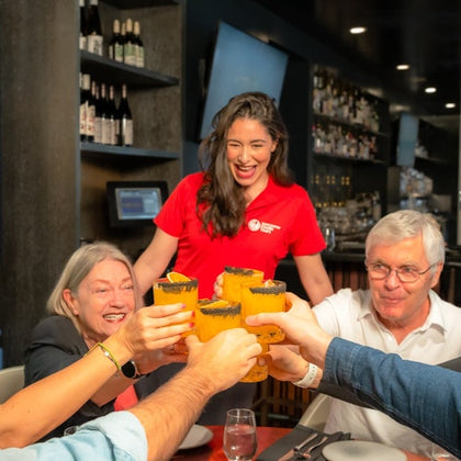 A group of people raising orange-colored drinks in a toast, with a smiling server in a red shirt in the background.