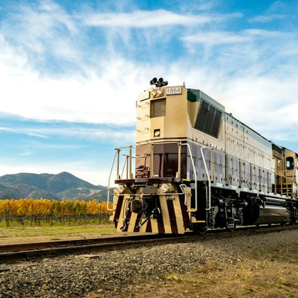 A beige locomotive traveling on tracks by a vineyard, with mountains and a blue sky in the background.