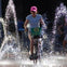 A person rides a bicycle through a plaza with water jets spraying around, wearing a pink cap and a light blue shirt.