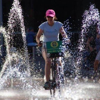 A person rides a bicycle through a plaza with water jets spraying around, wearing a pink cap and a light blue shirt.