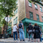 A group of people stands on a street corner, looking up at a two-story building with brick and wood siding in an urban setting.