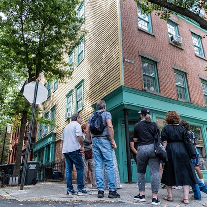 A group of people stands on a street corner, looking up at a two-story building with brick and wood siding in an urban setting.