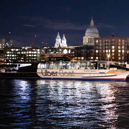 A boat labeled 'Uber Boat by Thames Clippers' docked on a river at night, with illuminated buildings and a dome in the background.