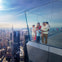 A family stands on a glass observation deck overlooking a city skyline on a sunny day.