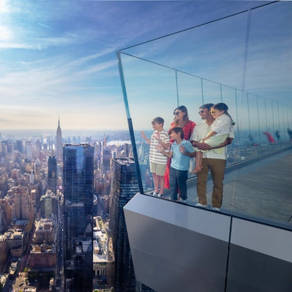 A family stands on a glass observation deck overlooking a city skyline on a sunny day.