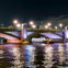 A night view of a bridge with decorative lighting over a river, with a boat passing underneath and a dome visible in the background.
