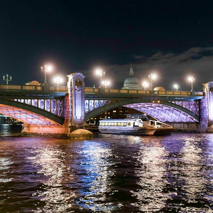 A night view of a bridge with decorative lighting over a river, with a boat passing underneath and a dome visible in the background.