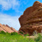 Red rock formations rise above a green field under a partly cloudy blue sky.