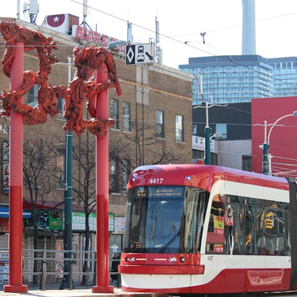 Red and white tram passing a street with red dragon sculptures on poles, against a backdrop of urban buildings and the CN Tower.