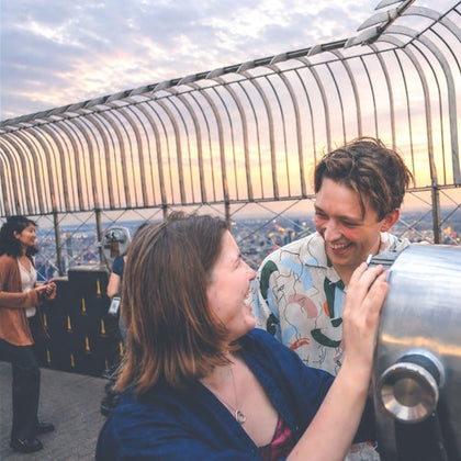 People enjoying the view from a fenced observation deck at sunset, with a cityscape visible in the background.
