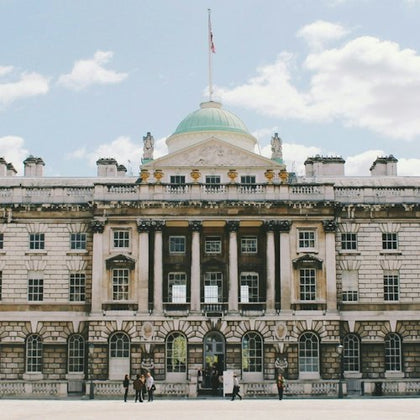 A neoclassical building with columns and a central dome, featuring a flag on top, with people in front on a sunny day.