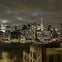 A city skyline at night with illuminated skyscrapers under a cloudy sky, reflecting on the water below.