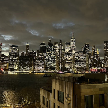 A city skyline at night with illuminated skyscrapers under a cloudy sky, reflecting on the water below.