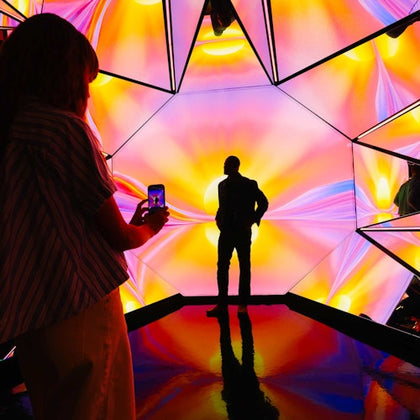 A person stands silhouetted against a colorful geometric light display, while another person photographs the scene.