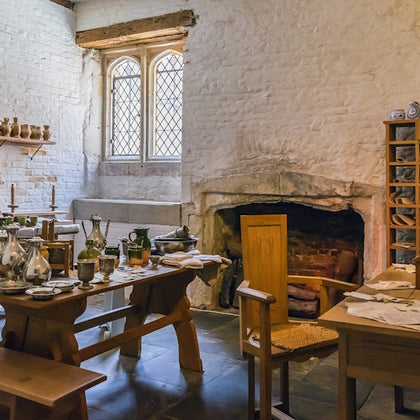 A rustic room with wooden furniture, a large fireplace, candles, glassware, and a multi-paned window.