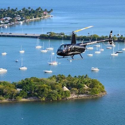 A black helicopter flying above a coastal area with sailboats on the water and an island with trees and huts visible below.