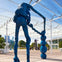 Blue abstract sculpture of a figure with a chain-like object, set in an outdoor space with a glass canopy and people in the background.