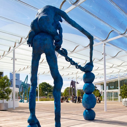 Blue abstract sculpture of a figure with a chain-like object, set in an outdoor space with a glass canopy and people in the background.