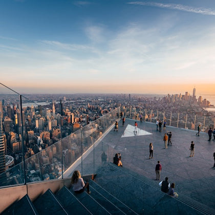 People on a rooftop observation deck with glass railings, overlooking a cityscape at sunset.