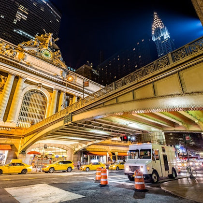 Night scene of Grand Central Terminal with yellow taxis and a white truck, under an overpass with the Chrysler Building in view.