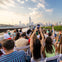A group of people seated on a boat, many holding up phones, with a city skyline and river in the background.