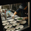 Chef in a kitchen plating dishes with greens, surrounded by bowls, utensils, and kitchen equipment.