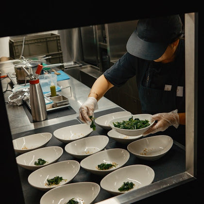 Chef in a kitchen plating dishes with greens, surrounded by bowls, utensils, and kitchen equipment.