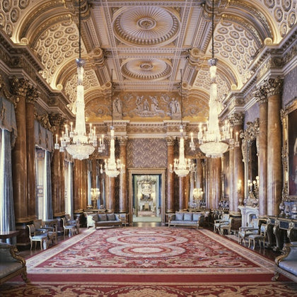 Ornate grand hall with chandeliers, detailed ceiling, marble columns, and red carpet. Furniture lines the sides, with large mirrors and paintings.