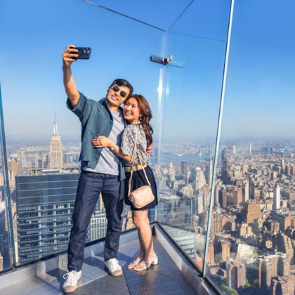 A couple taking a selfie on a glass observation deck overlooking a cityscape with the Empire State Building visible.