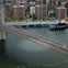 A helicopter flying near a bridge over a river, with city buildings and a roadway with cars in the background.