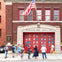 People standing in front of a fire station with red doors and an American flag flying above. The building is labeled "ENGINE 205 H&L 118."