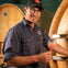 A man in glasses and a cap holds a glass of pink liquid, standing in front of large wooden barrels.