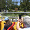 Tourists on the upper deck of an open-top bus photographing a 'Beverly Hills' sign in a green park setting.