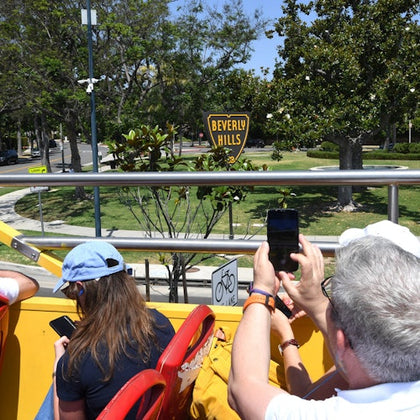 Tourists on the upper deck of an open-top bus photographing a 'Beverly Hills' sign in a green park setting.
