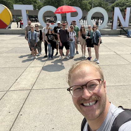 A group of people posing in front of large "Toronto" letters in a plaza, with one person in the foreground taking a selfie.
