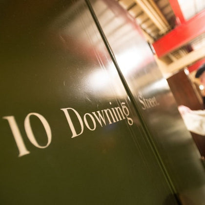 Close-up of a green door with "10 Downing Street" written on it in white letters, indoors with red structural beams visible.